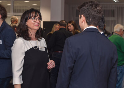 Eine Frau mit Brille und Perlenkette unterhält sich mit einem Mann in Anzug. Im Hintergrund sind weitere Personen bei einem Event zu sehen.