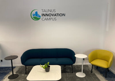 Ein moderner Lounge-Bereich mit einem dunkelblauen Sofa, einem gelben Sessel, Beistelltischen und einer kleinen Pflanze darauf. Im Hintergrund ist das Logo des "Taunus Innovation Campus" an der Wand sichtbar.