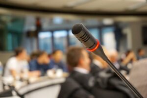 Ein schwarzes Mikrofon mit rotem Leuchtring im Fokus, im Hintergrund unscharf eine Gruppe von Menschen in einem Konferenzraum.