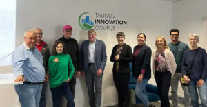 Gruppe von Personen beim Treffen im Taunus Innovation Campus vor dem TIC-Logo in modernen Büroräumen.